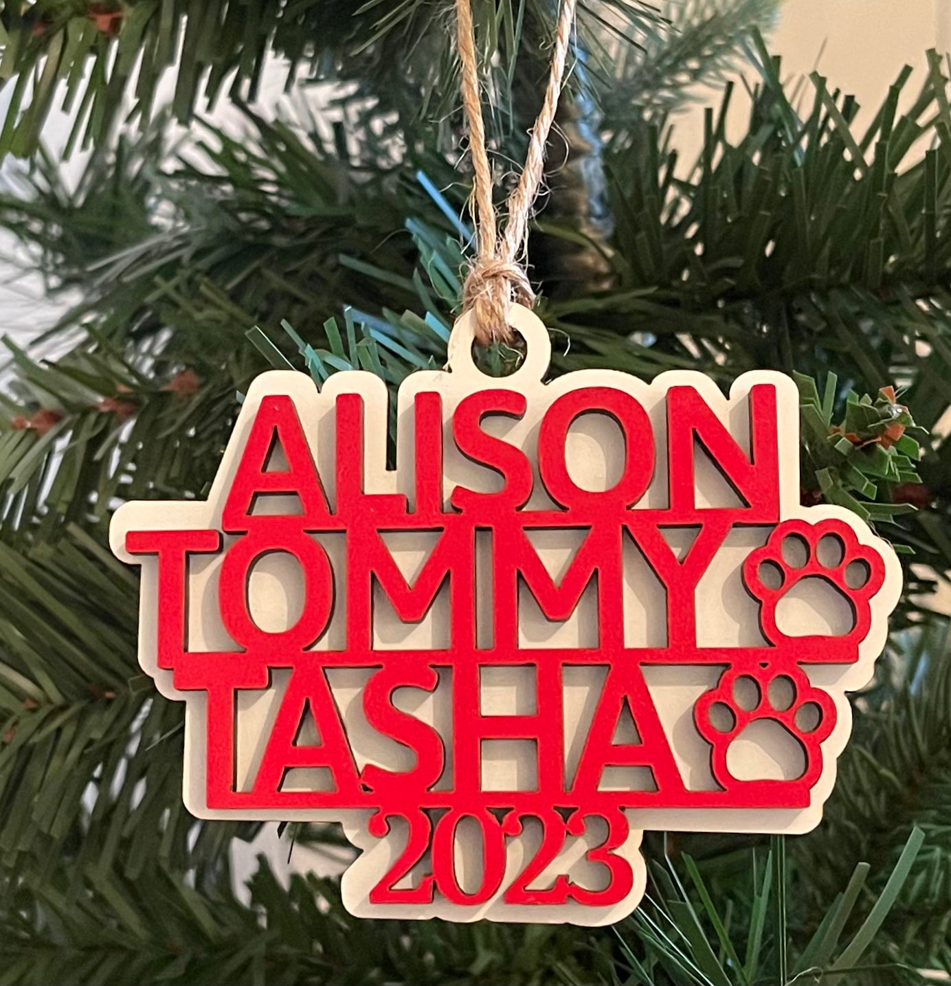 A name and two pet names are shown on the custom ornament, with pet paws, the year of 2023, and a white backer with jute twine hanger for the ornament.