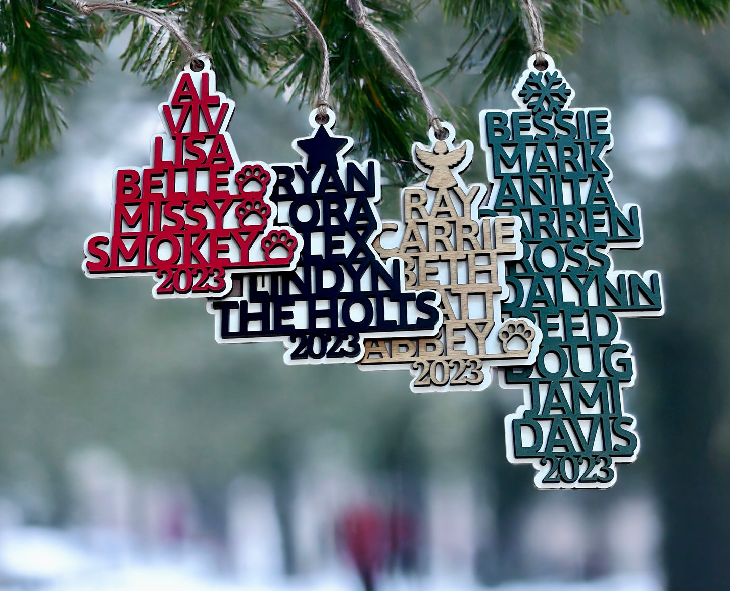 Examples of Art by Iceberg's personalized name family Christmas tree ornaments are shown hanging from a green tree, with a snowy scene in the background.