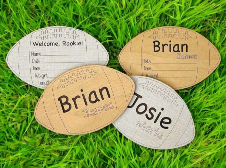 Football-shaped baby birth announcement sign. Newborn gift. Welcome, rookie is engraved, and below is an area to fill in the baby name and birth stats. Choose linen or maple wood color option. From Art by Iceberg.