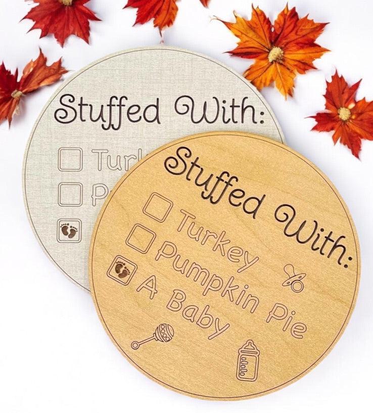 Fall Thanksgiving family gathering baby pregnancy surprise announcement sign. Five inches round, choose linen or maple wood. Engraved wording says: Stuffed With and check-mark boxes for turkey, pumpkin pie, or a baby. Baby box has baby feet in it.